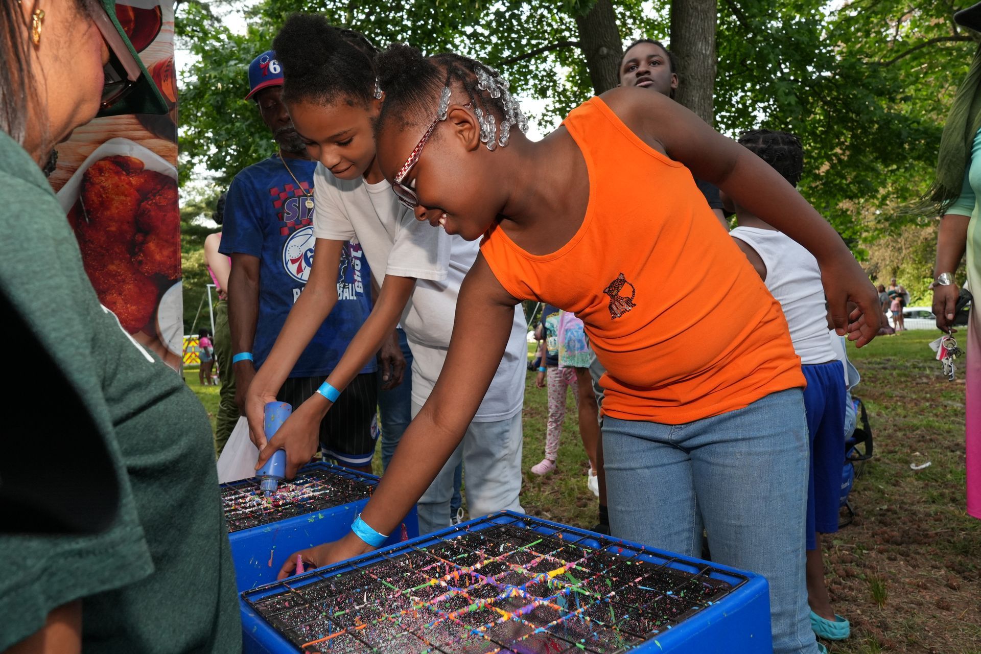 Two people in orange and white shirts lean over a blue bin at an outdoor table, sorting colorful beads in a park