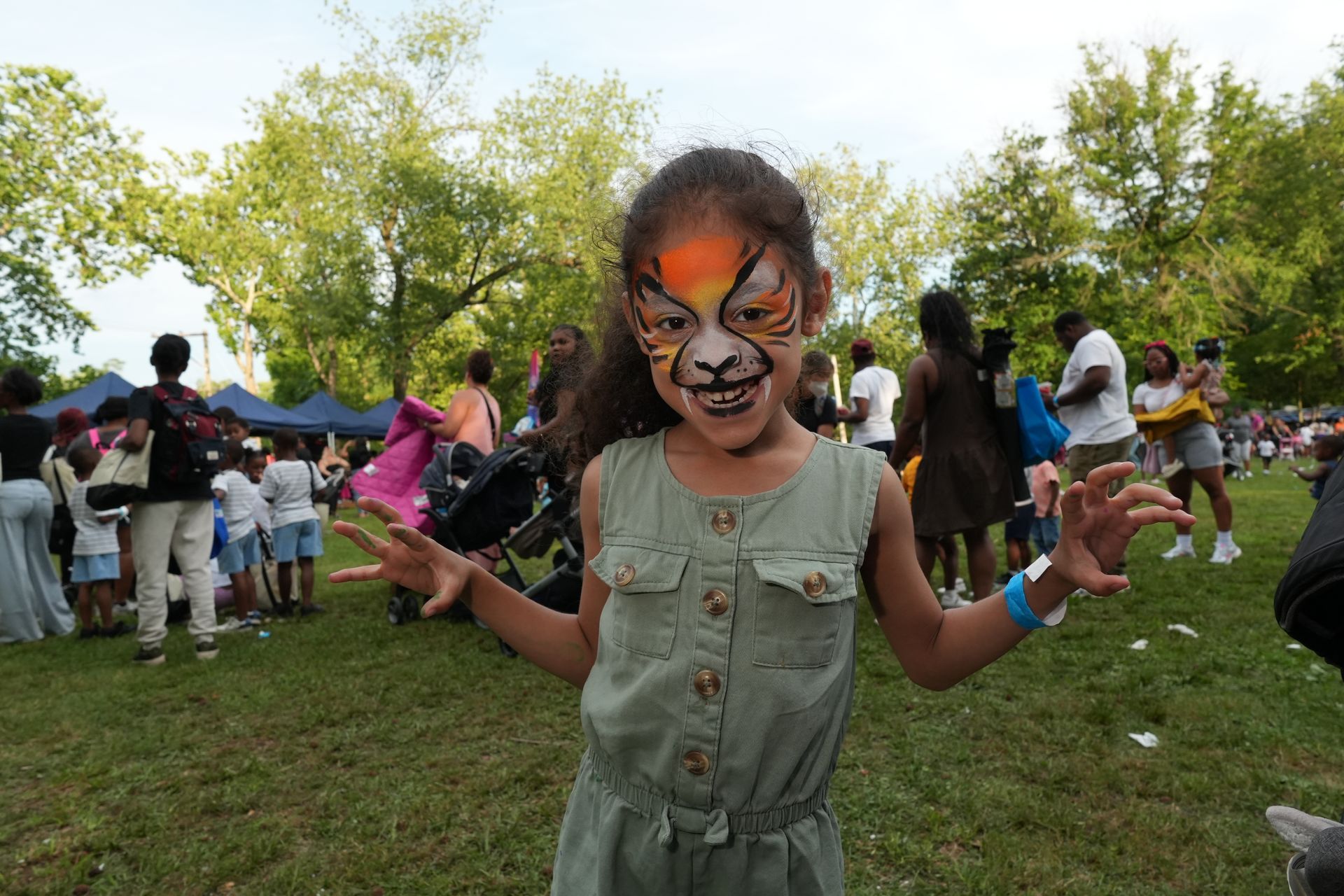 Child with tiger face paint stands in a park crowd, wearing a green dress and smiling with raised hands.