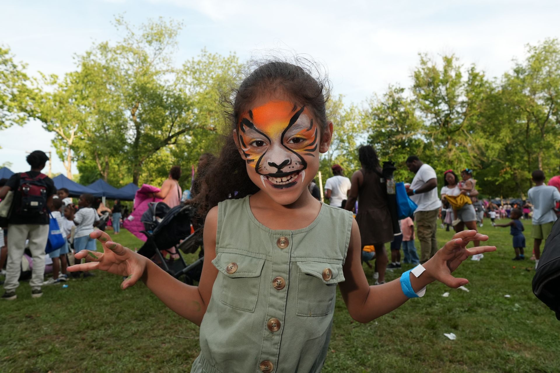 Child with tiger face paint and outstretched hands at an outdoor festival in a grassy park