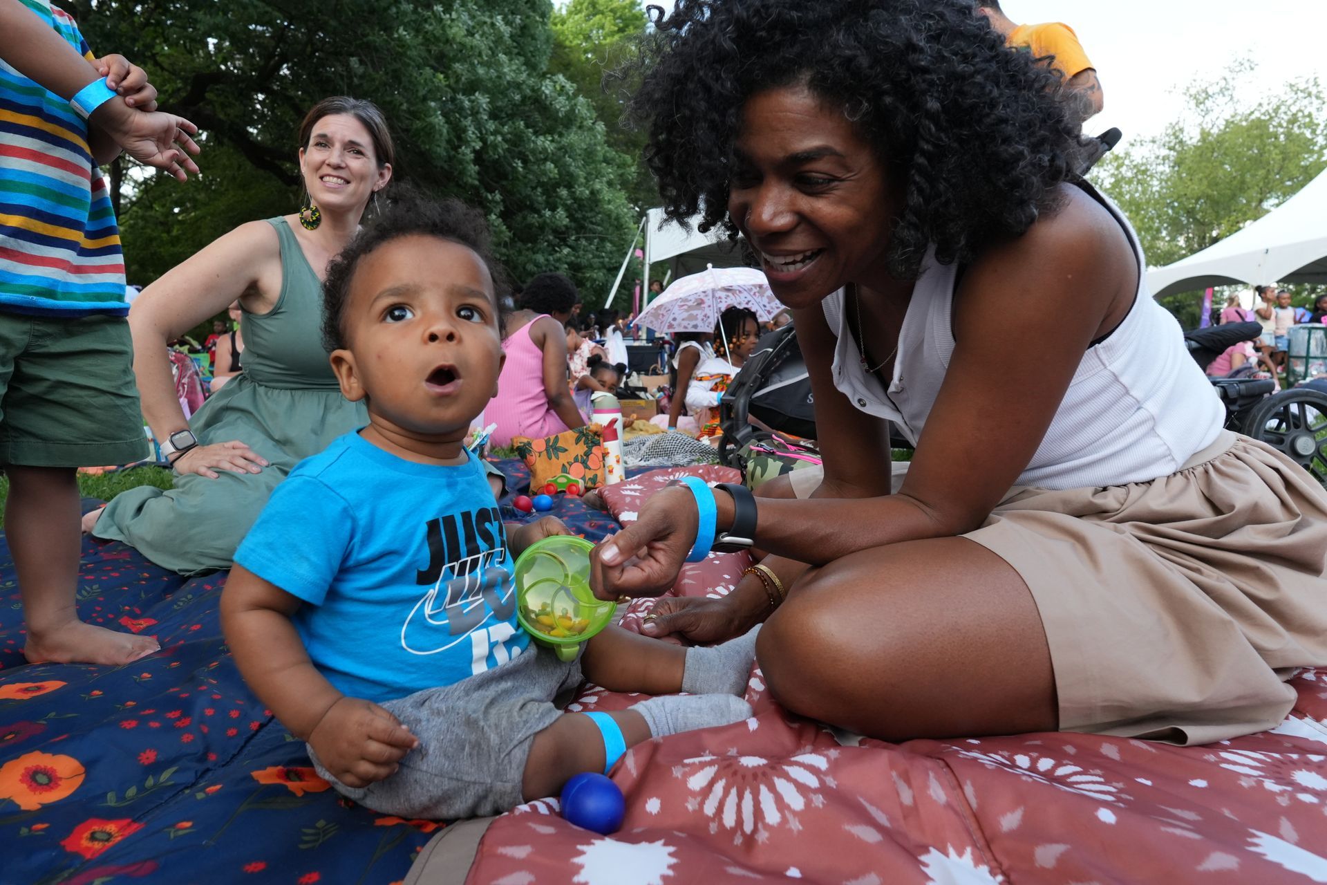 Adult and toddler on patterned blanket at outdoor gathering, with other people and tents in background
