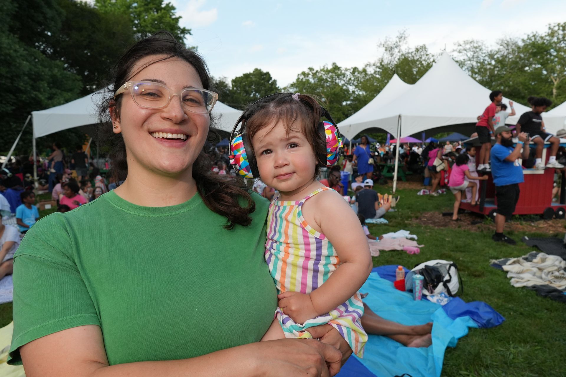 Smiling woman holding a child at an outdoor event with tents and people in the background