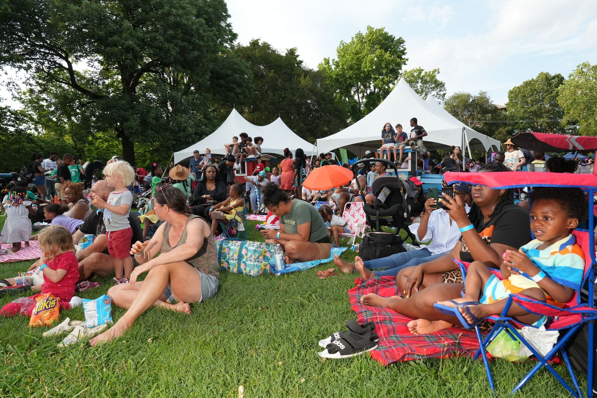 Crowd seated on blankets and chairs at an outdoor festival under white tents on a grassy field