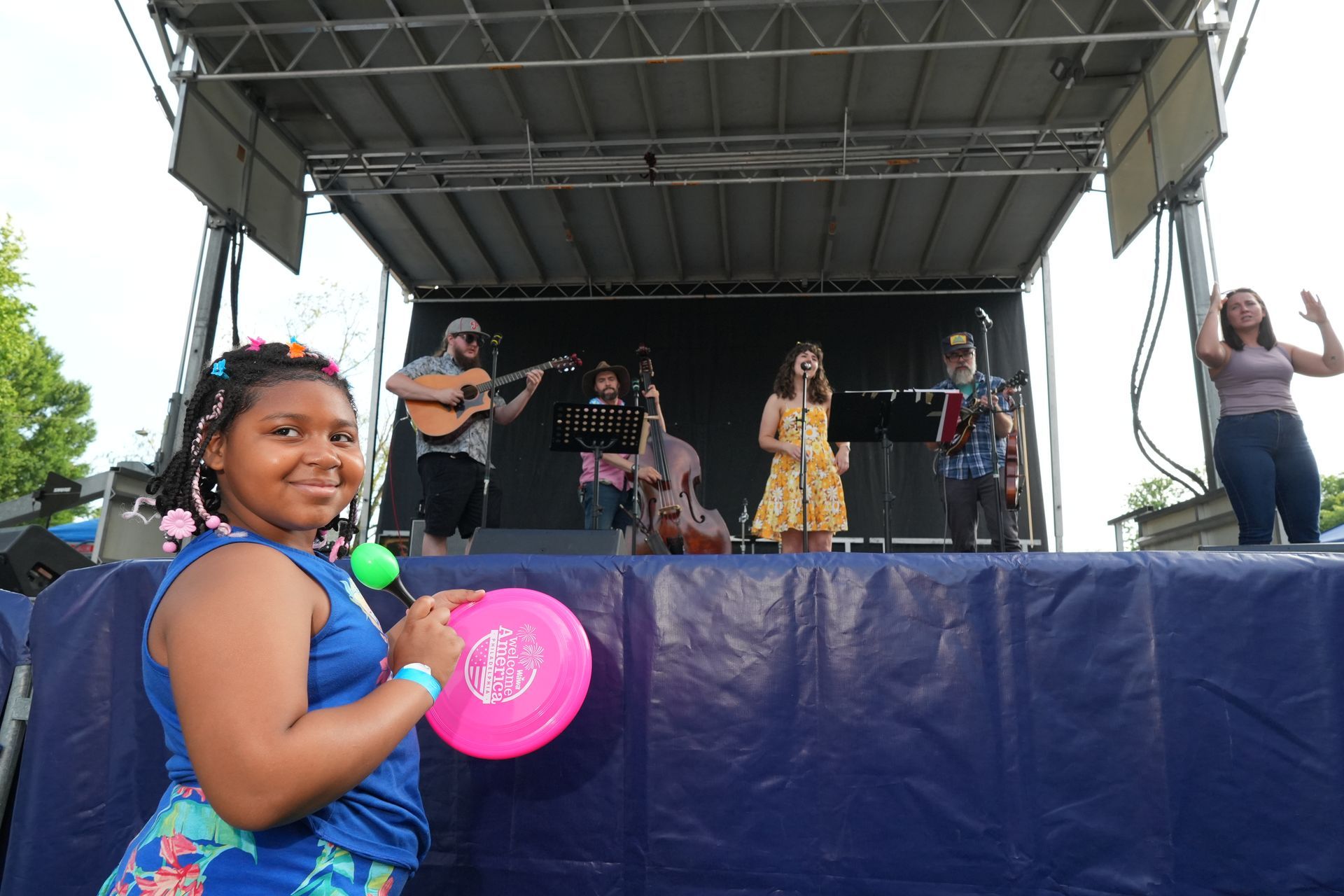 Children performing on an outdoor stage, with a smiling girl holding a pink fan in the foreground.