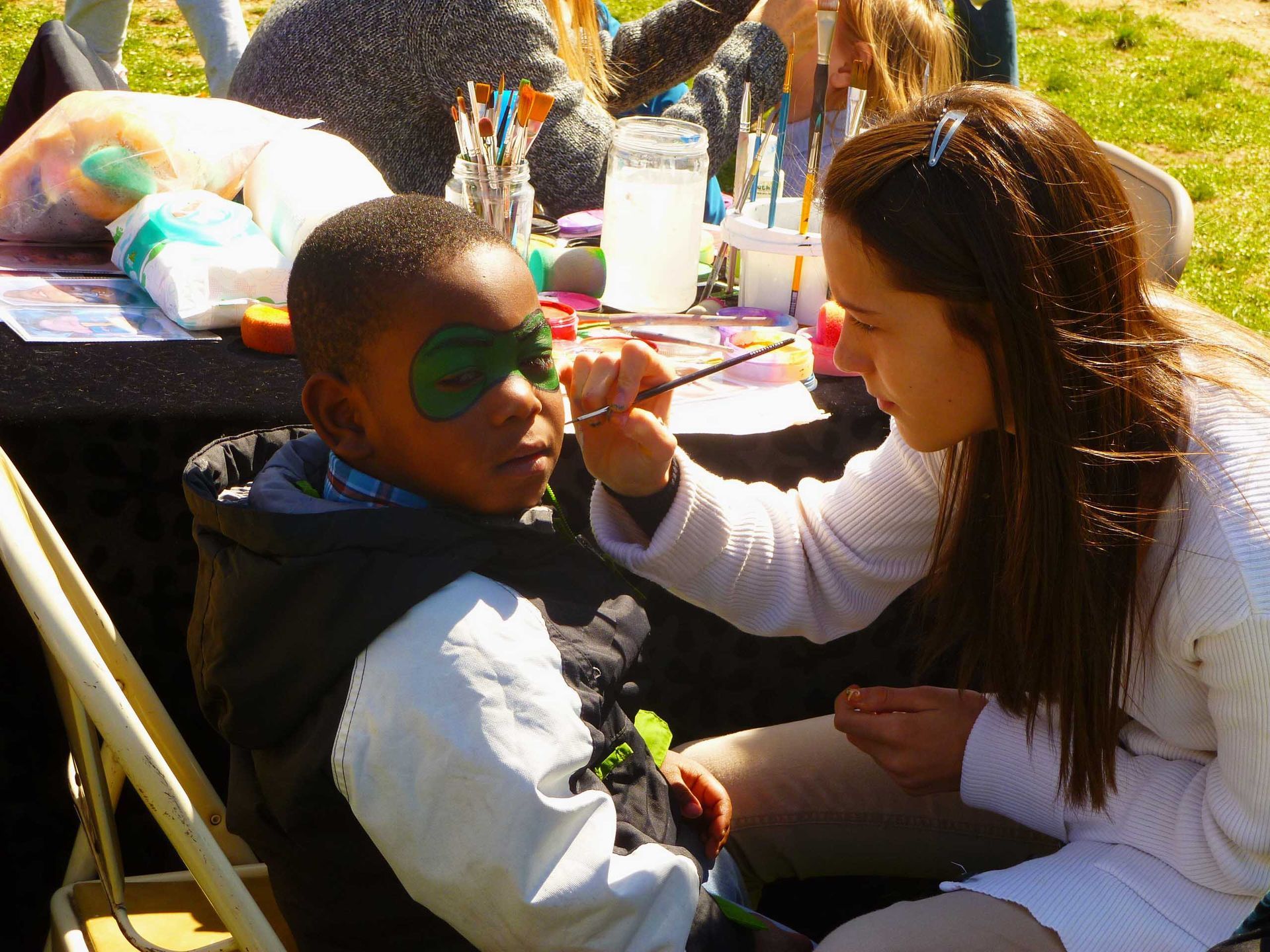 Woman paints a child's face green at an outdoor event table with art supplies.