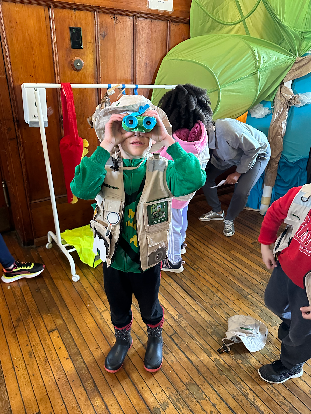 Child looking through toy binoculars in a wood-floored room with coats and backpacks hanging nearby