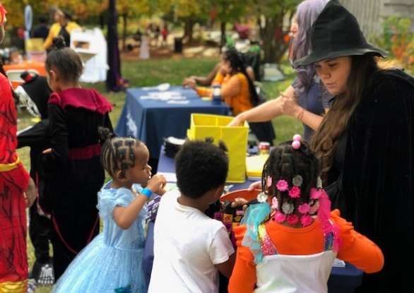 Children at an outdoor event play with colorful toys beside a table with a woman in a black hat
