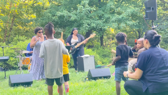 Outdoor live music performance in a grassy park with a singer, guitarist, and seated audience.