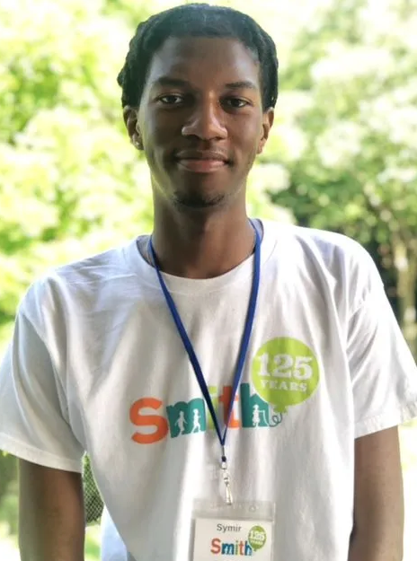 Smiling person wearing a white T-shirt and lanyard, standing outdoors with greenery in the background
