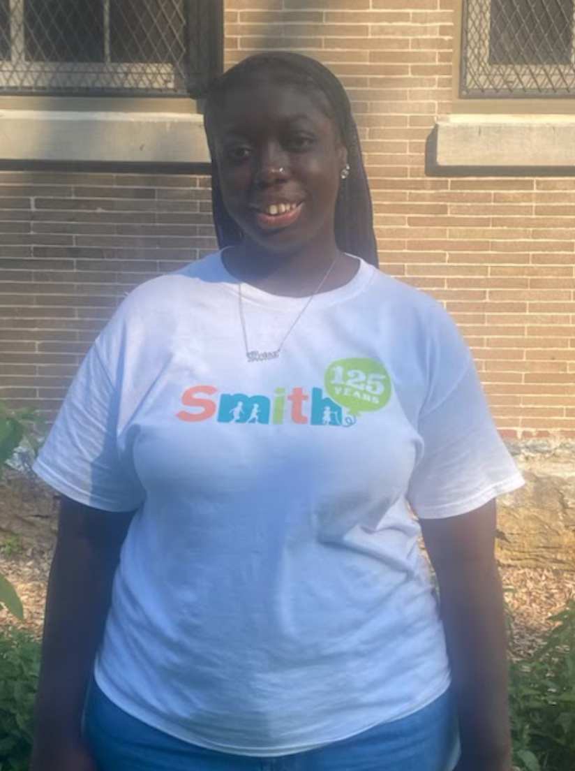 Person standing outdoors in a white “Smith” T-shirt, smiling in front of a brick wall.