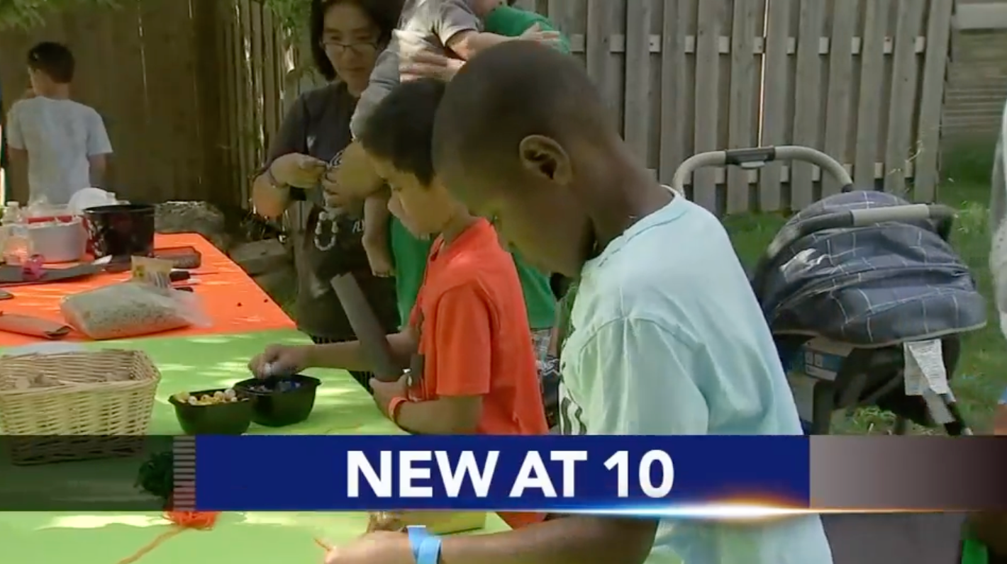 People gathered at a table preparing food or crafts outdoors; banner reads “NEW AT 10.”