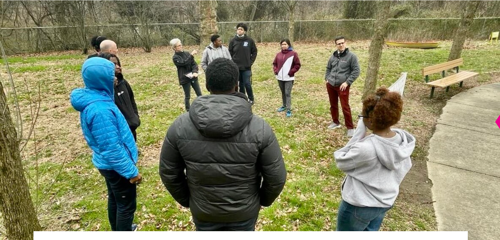 People standing in a wooded park, facing a guide during an outdoor group activity.