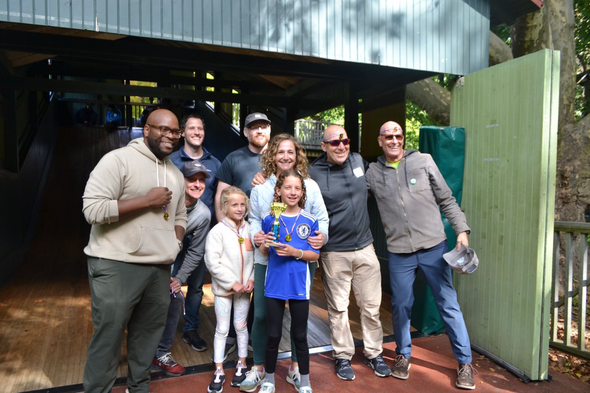 Group posing outdoors in front of a building, smiling and holding a blue stuffed toy.