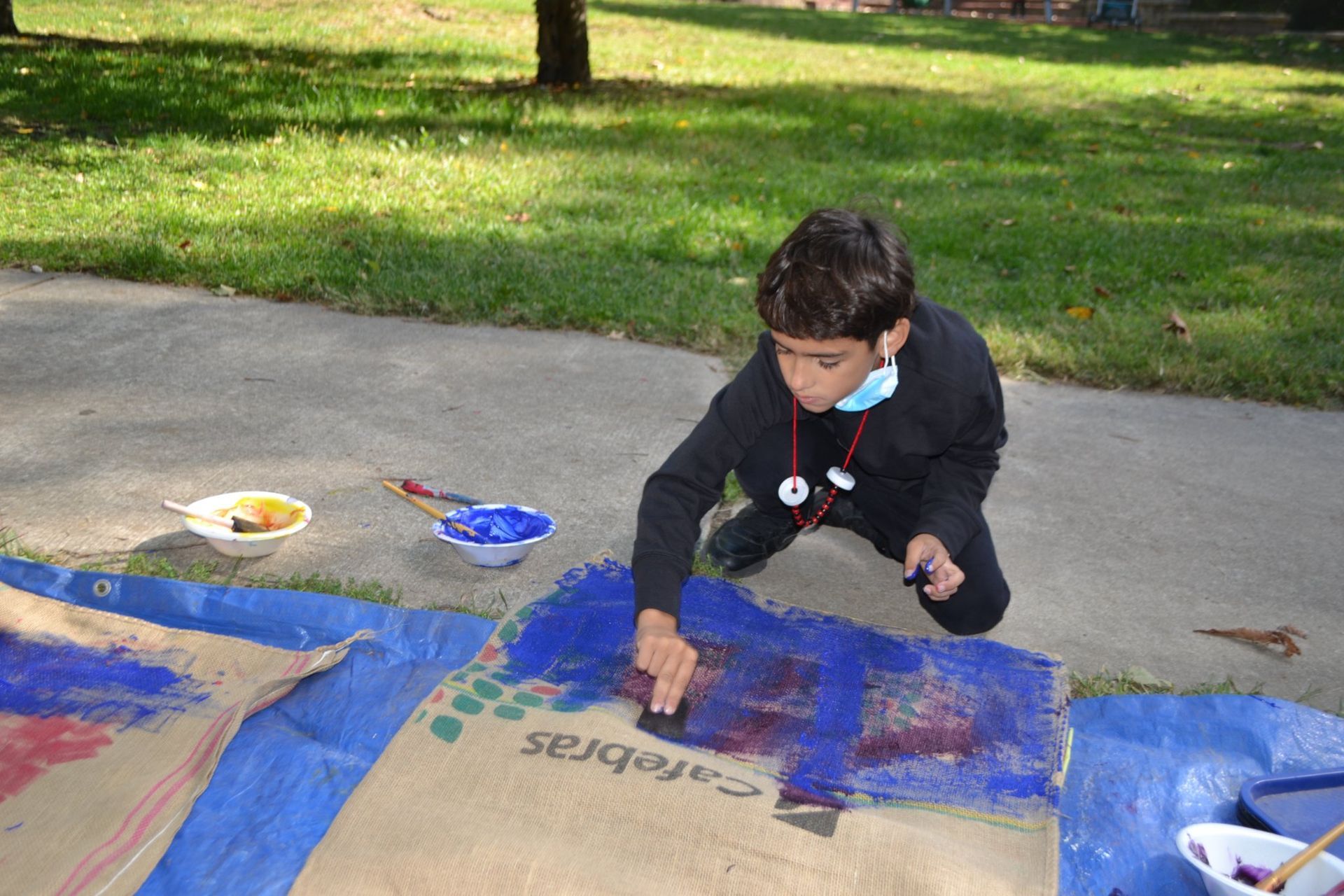 Child painting on cardboard outdoors with blue and red paint, beside paint trays on a concrete path.