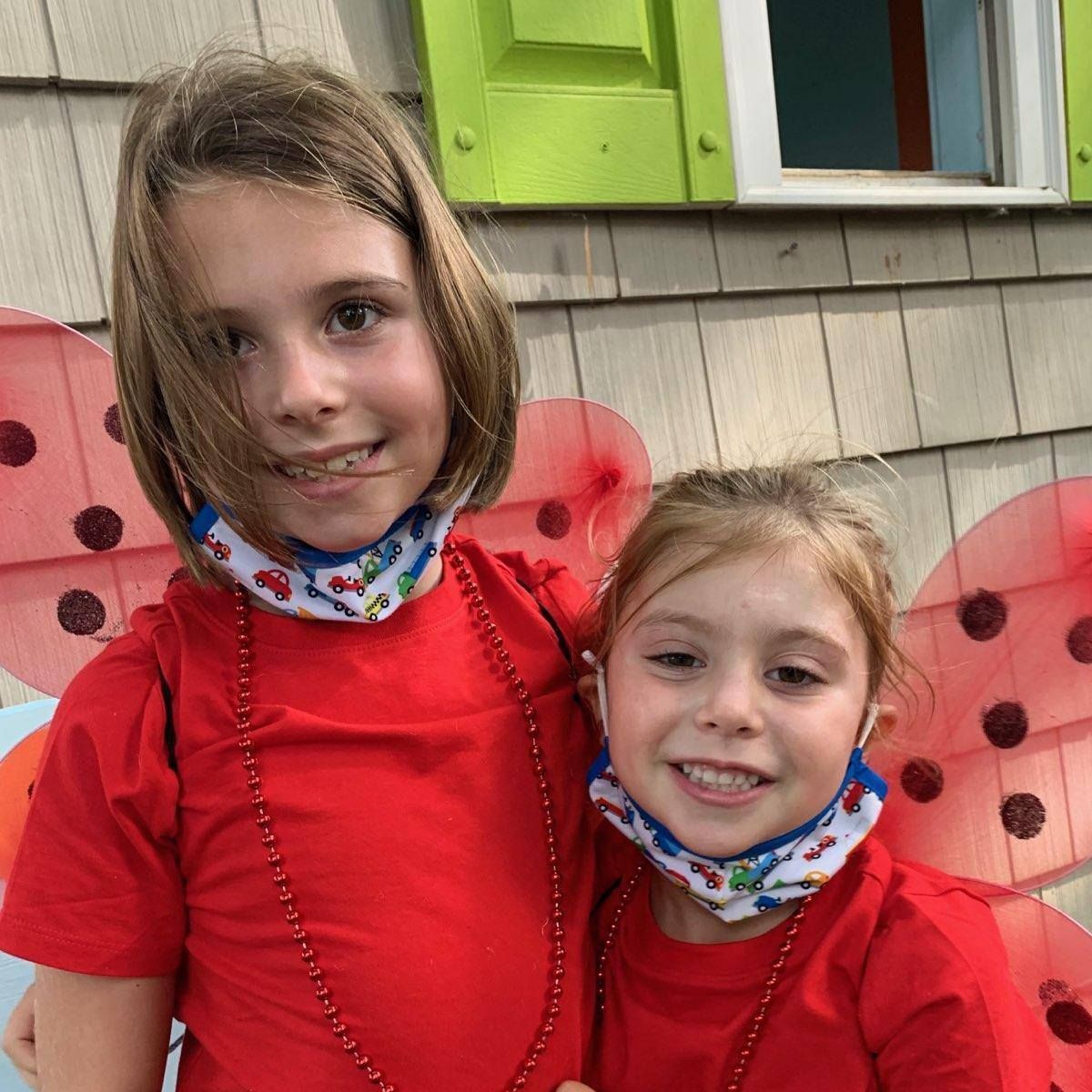 Two children in red shirts stand smiling in front of a house with butterfly wings behind them.