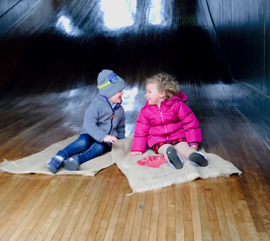 Two children in winter clothes sit on a rug on a wooden slide, facing each other and smiling.