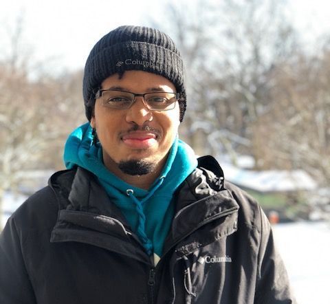 Man wearing glasses and a knit beanie outdoors in a black jacket and teal hoodie, standing in snowy sunlight