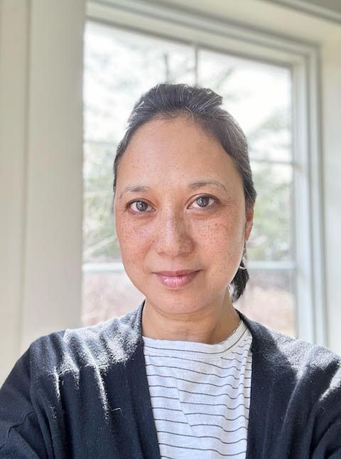 Portrait of a person indoors by a bright window, wearing a dark cardigan and striped shirt, with a slight smile