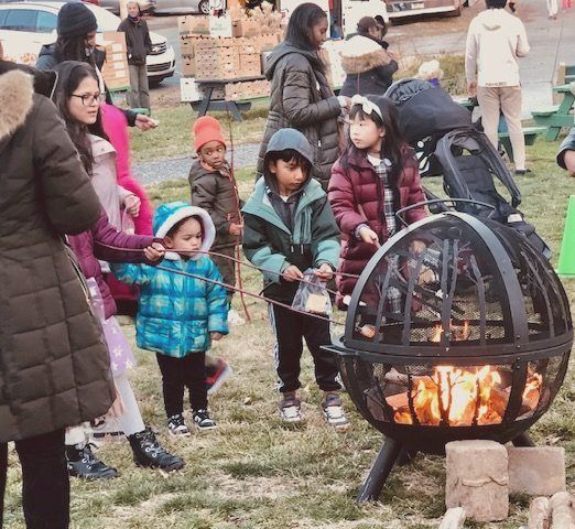 People gathered around a small outdoor fire pit on a grassy field at a community event.