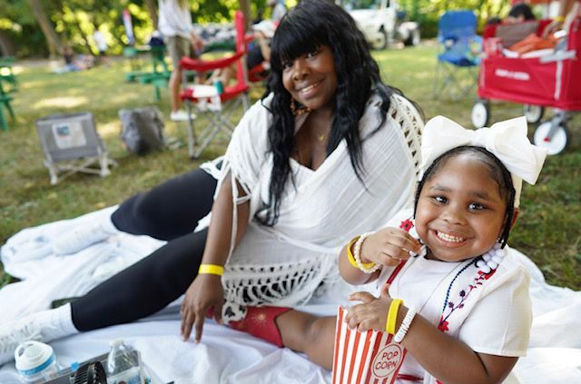 Two people smiling on a picnic blanket outdoors, sharing a snack in a park.