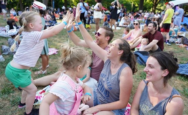 People sitting on grass at a lively outdoor festival, smiling and raising hands to a child standing nearby.