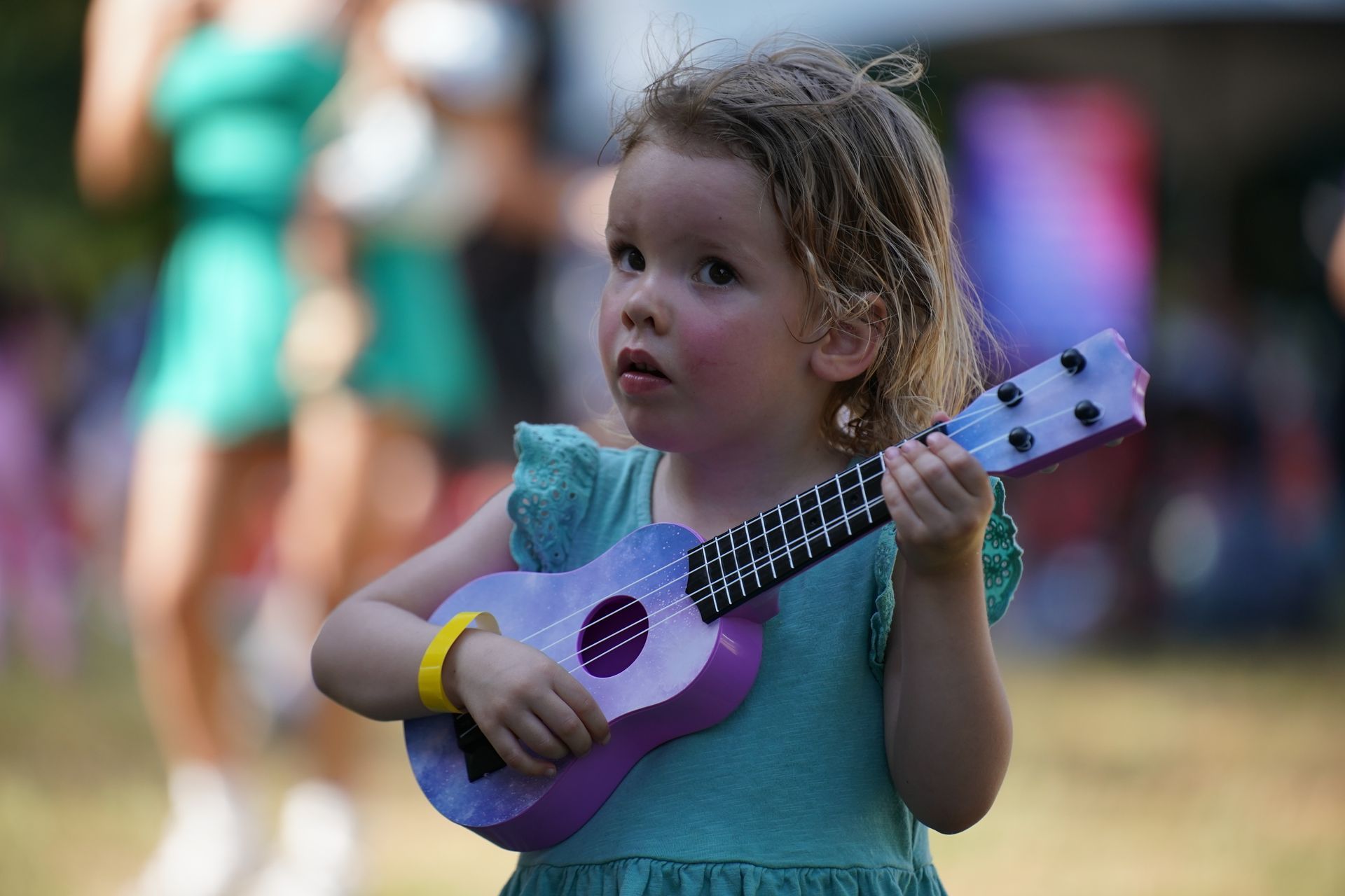 Child holding a purple ukulele outdoors, with a blurred crowd in the background