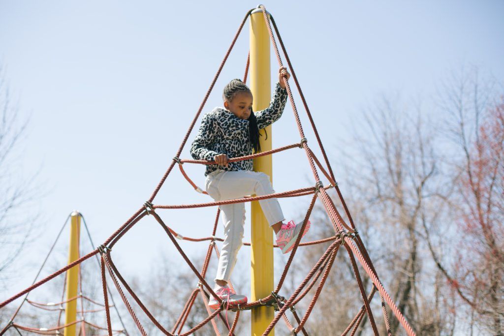 Person climbing a rope net playground structure with yellow posts outdoors against a blue sky