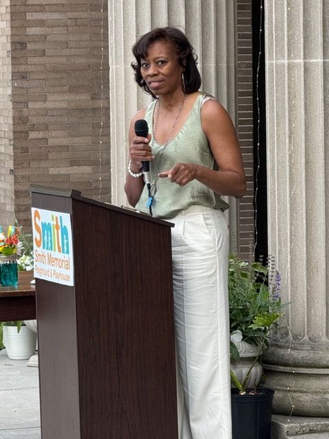 Woman speaking at a podium with a microphone in an outdoor courtyard.