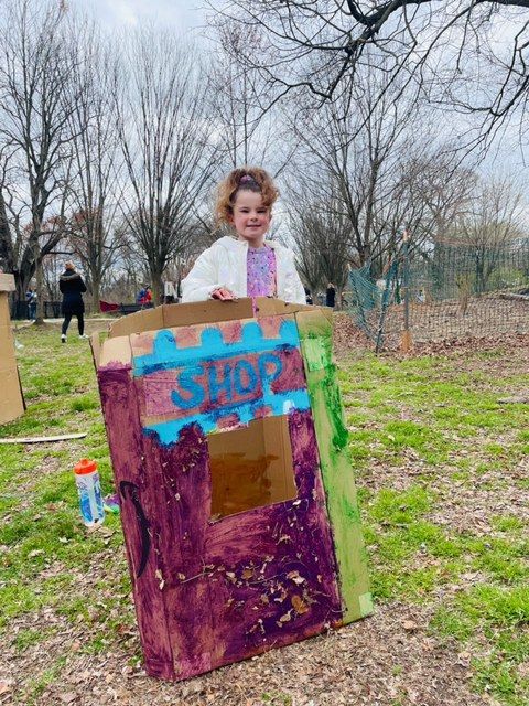 Child standing behind a colorful painted cardboard play booth in a park, with bare trees and grass.
