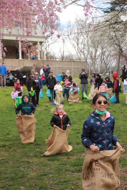 Children in burlap sacks racing on a grassy field with spectators and trees in the background