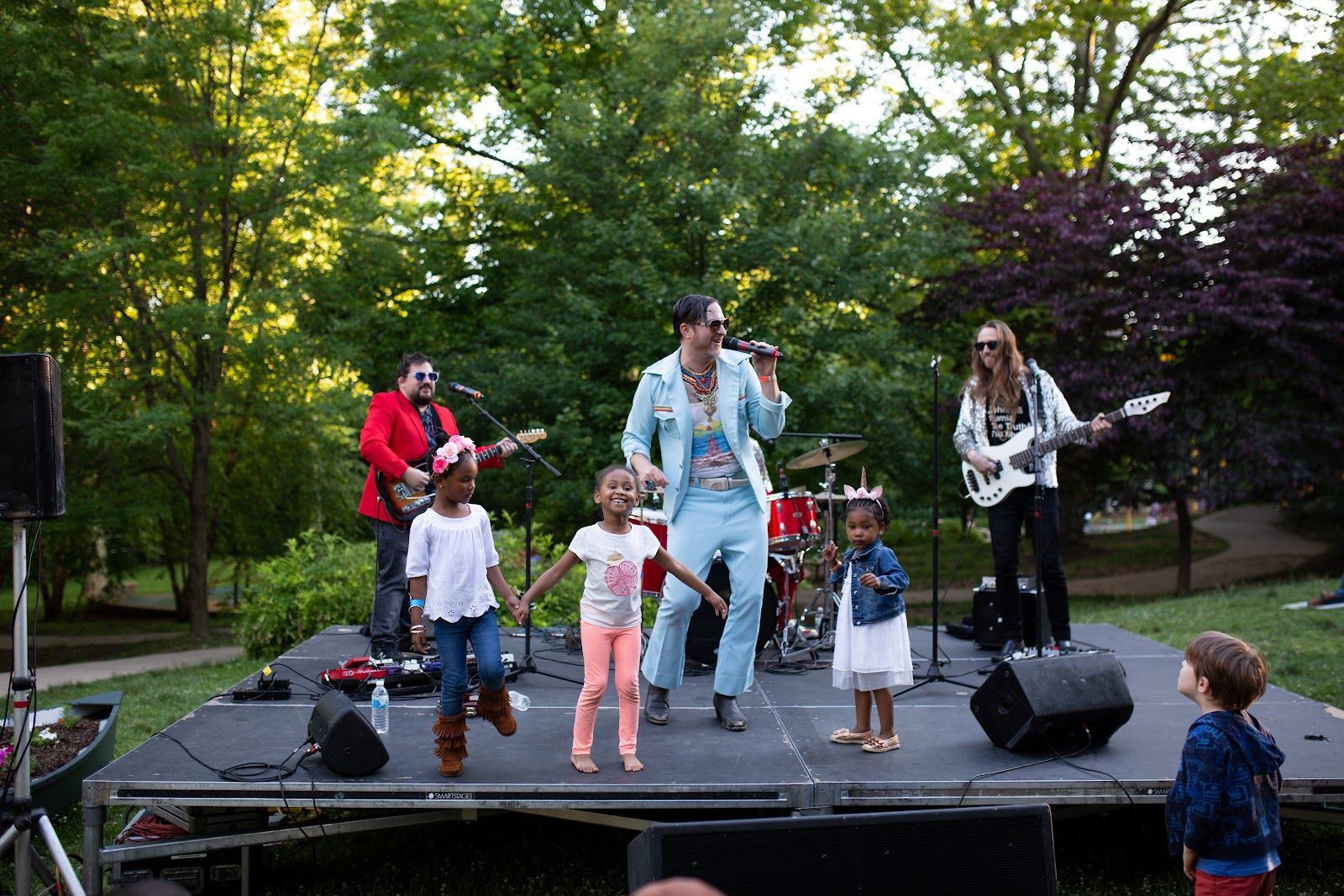 Band performing outdoors on a small stage with children dancing in front, surrounded by trees.