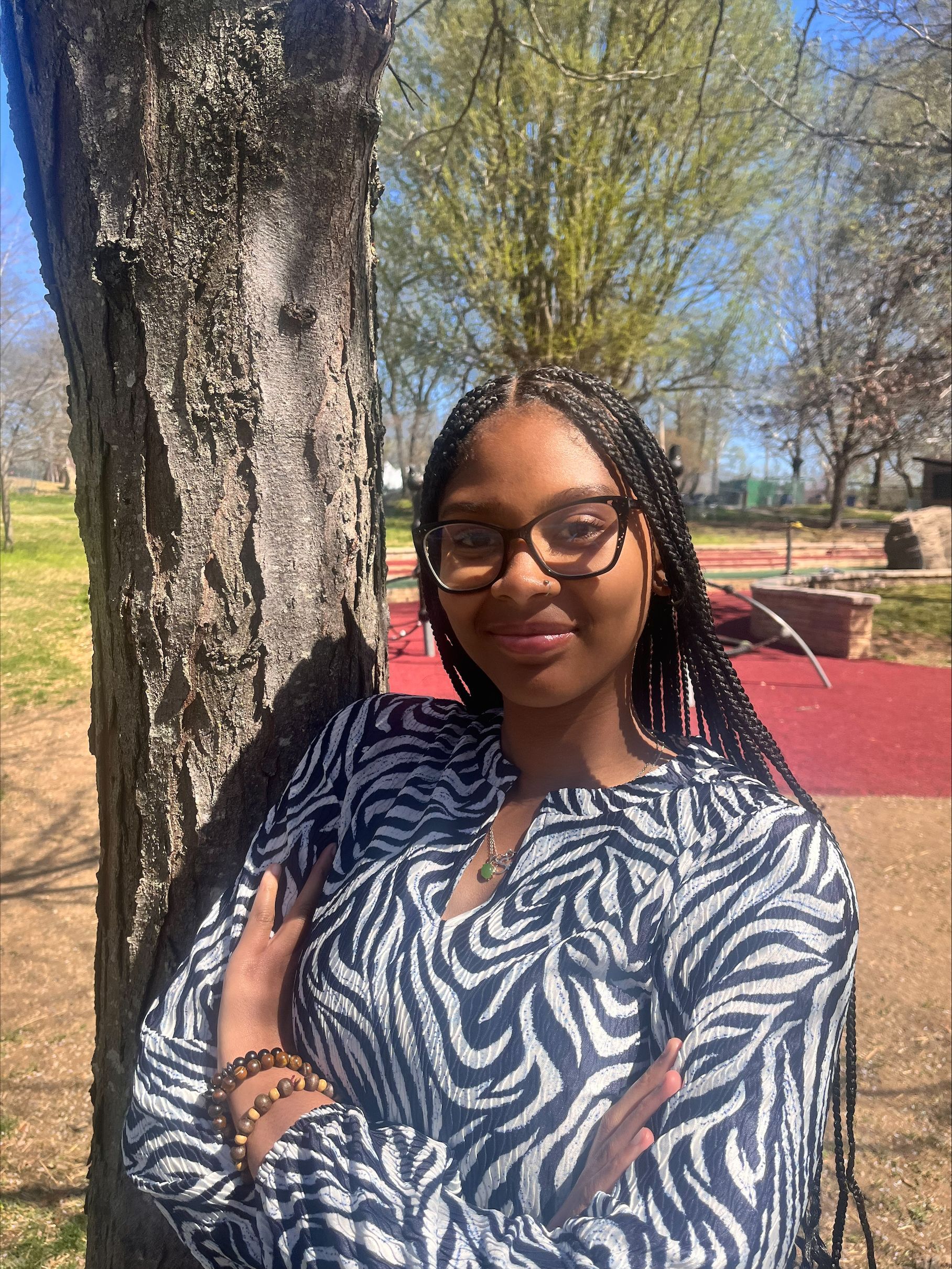 Woman leaning against a tree outdoors, wearing glasses and a blue patterned shirt, with a slight smile.