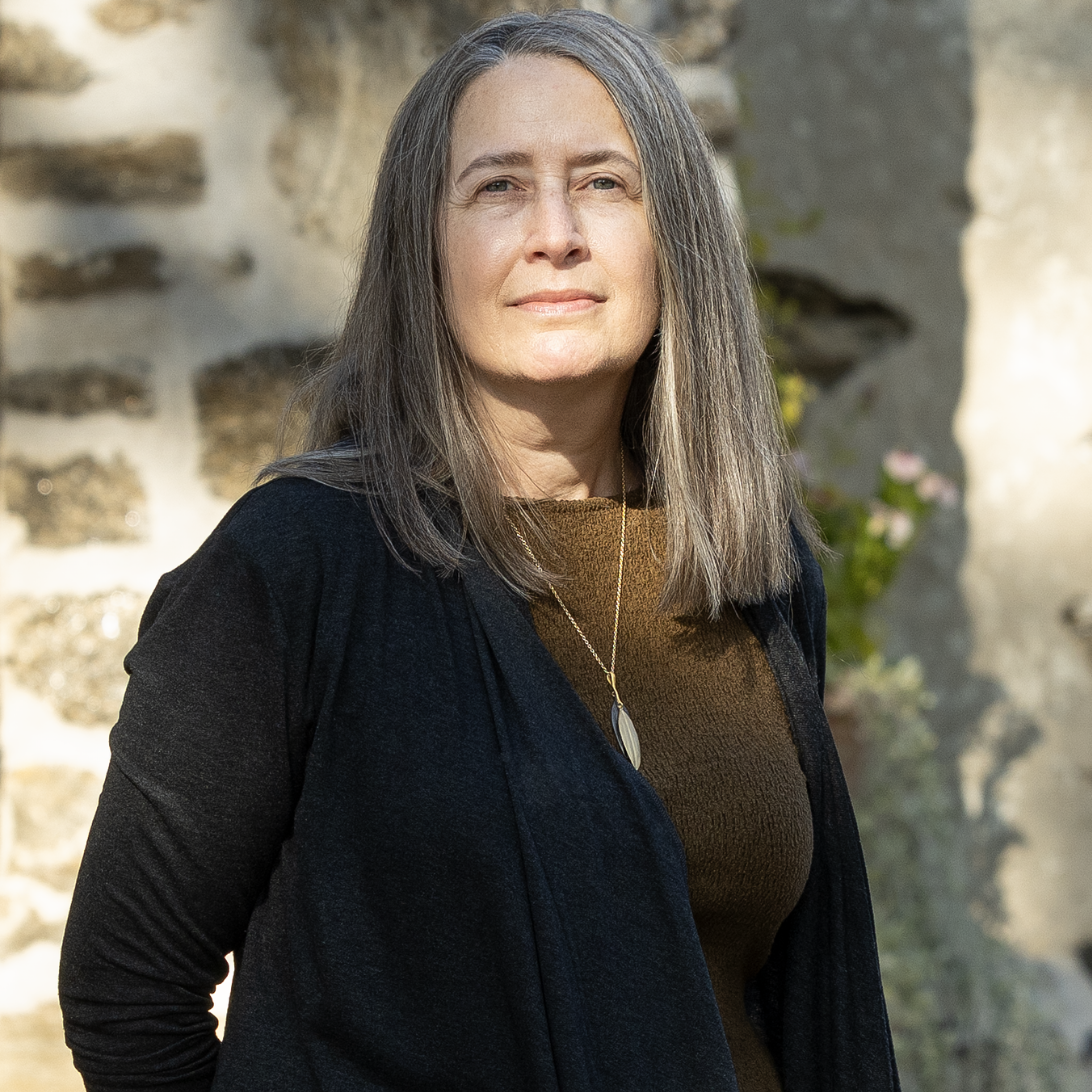 Woman in a black sweater standing outdoors against a stone wall in sunlight.
