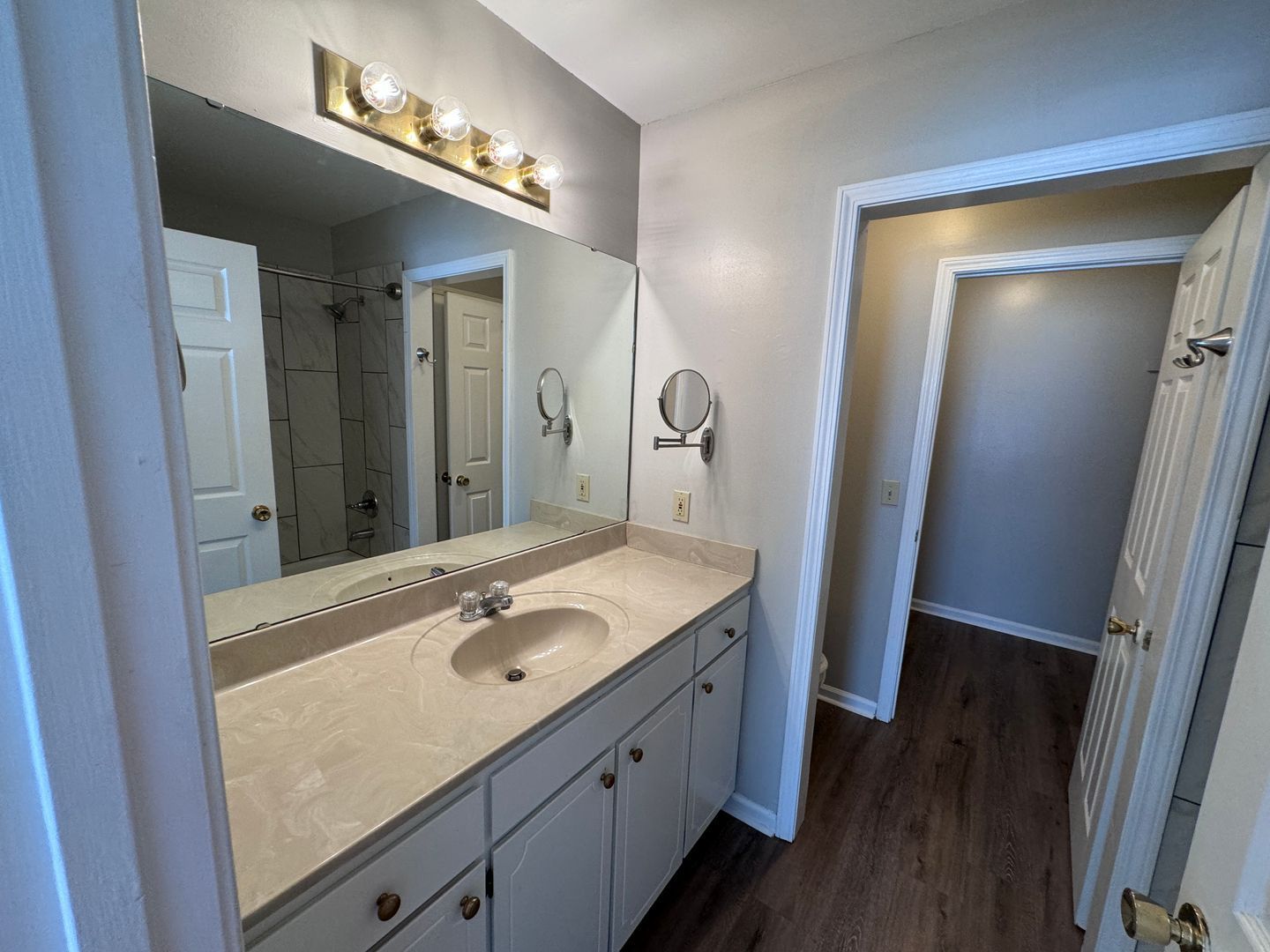 Bathroom with a vanity, mirror, and doorway. Light-colored countertop, white cabinets, and wood-look flooring.