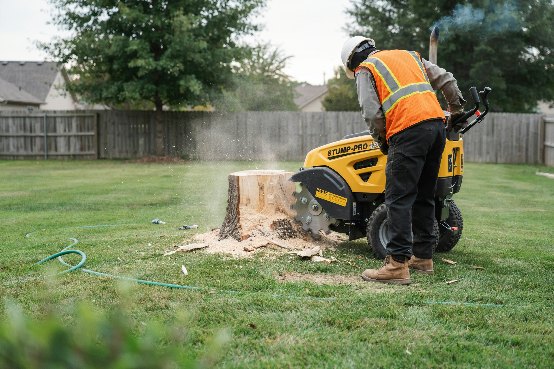 Worker in orange vest operating a yellow stump grinder in a backyard, spraying wood chips