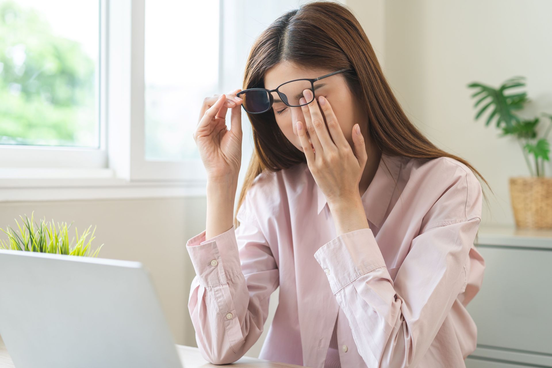 Person adjusting glasses while sitting at a desk with a laptop near a bright window. Person adjusting glasses while sitting at a desk with a laptop near a bright window.