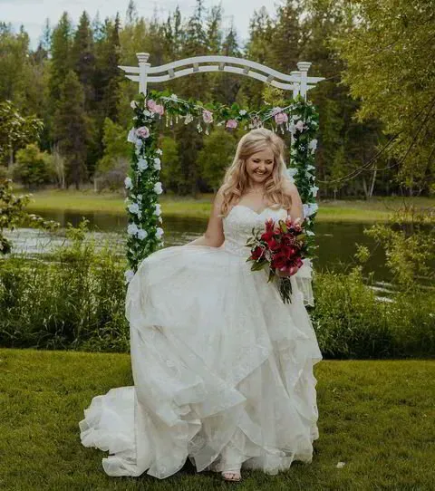 Bride in a white gown holding a red bouquet by a floral arch beside a pond
