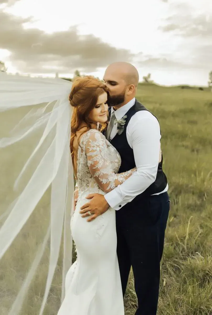 Couple embracing in a grassy field, bride in white dress and veil, groom in navy suit at sunset