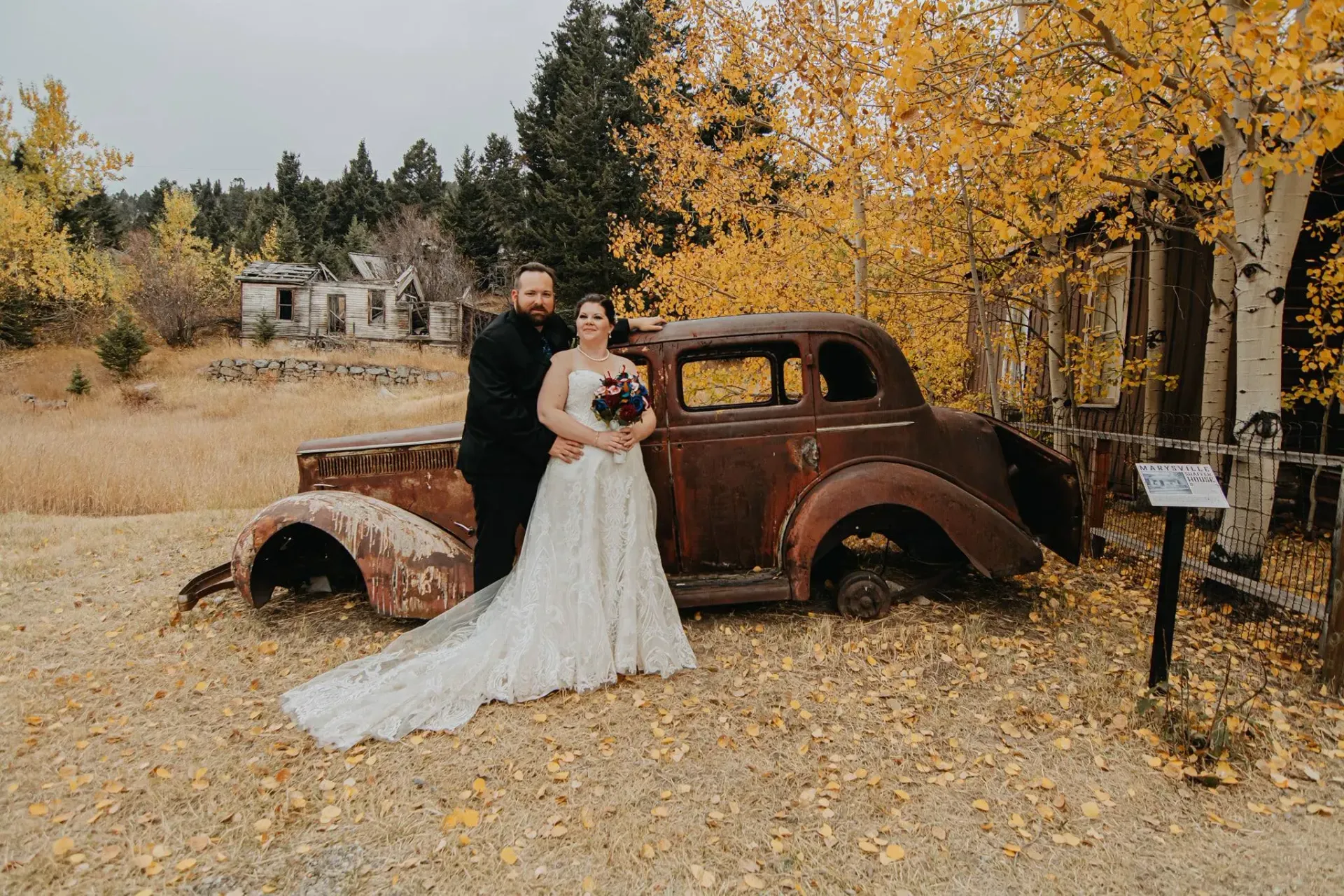 Bride and groom in white and black standing by a rusted vintage car under autumn trees.