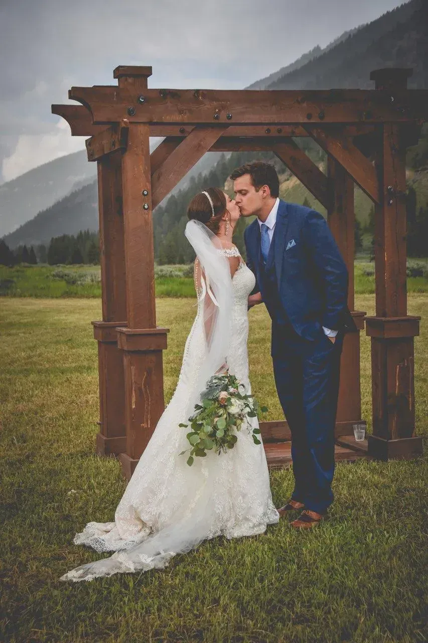 Bride and groom kiss under a wooden arch in a rainy mountain field.