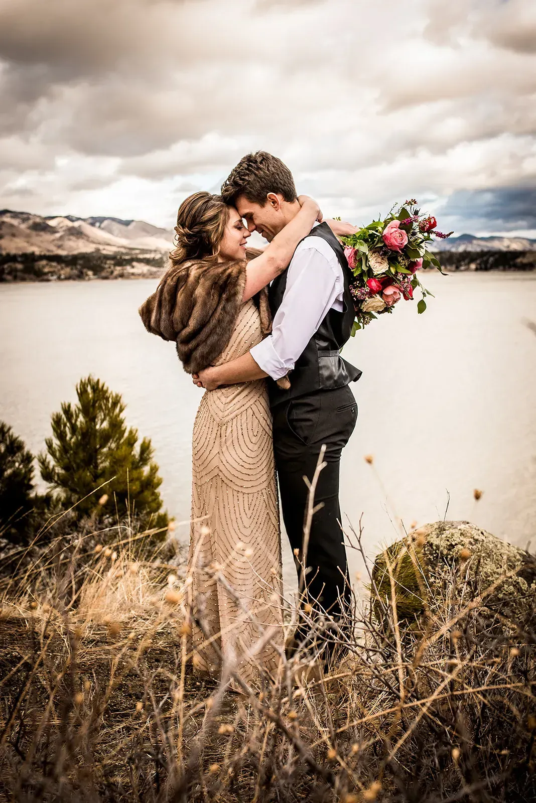Couple embracing outdoors by a lake, with mountains and cloudy sky behind them, holding a bouquet