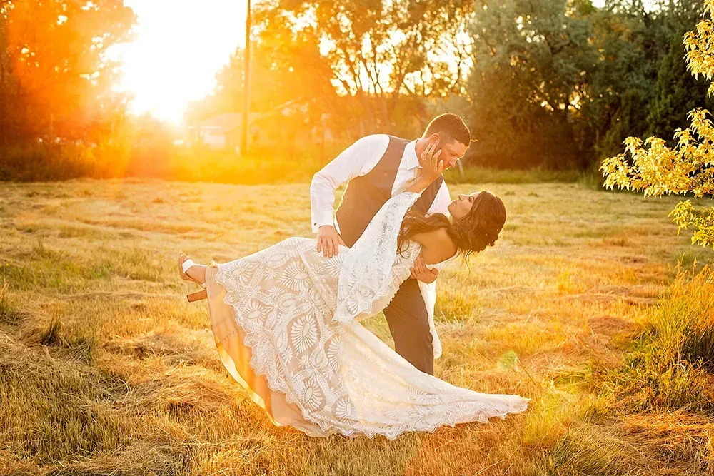 Couple dancing in a sunlit field, bride in white dress leaning back as groom holds her.