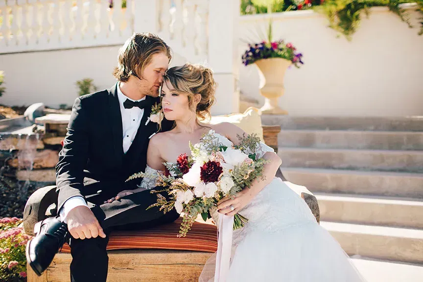 Newlywed couple sitting on a bench outdoors, sharing a kiss with a bouquet beside them