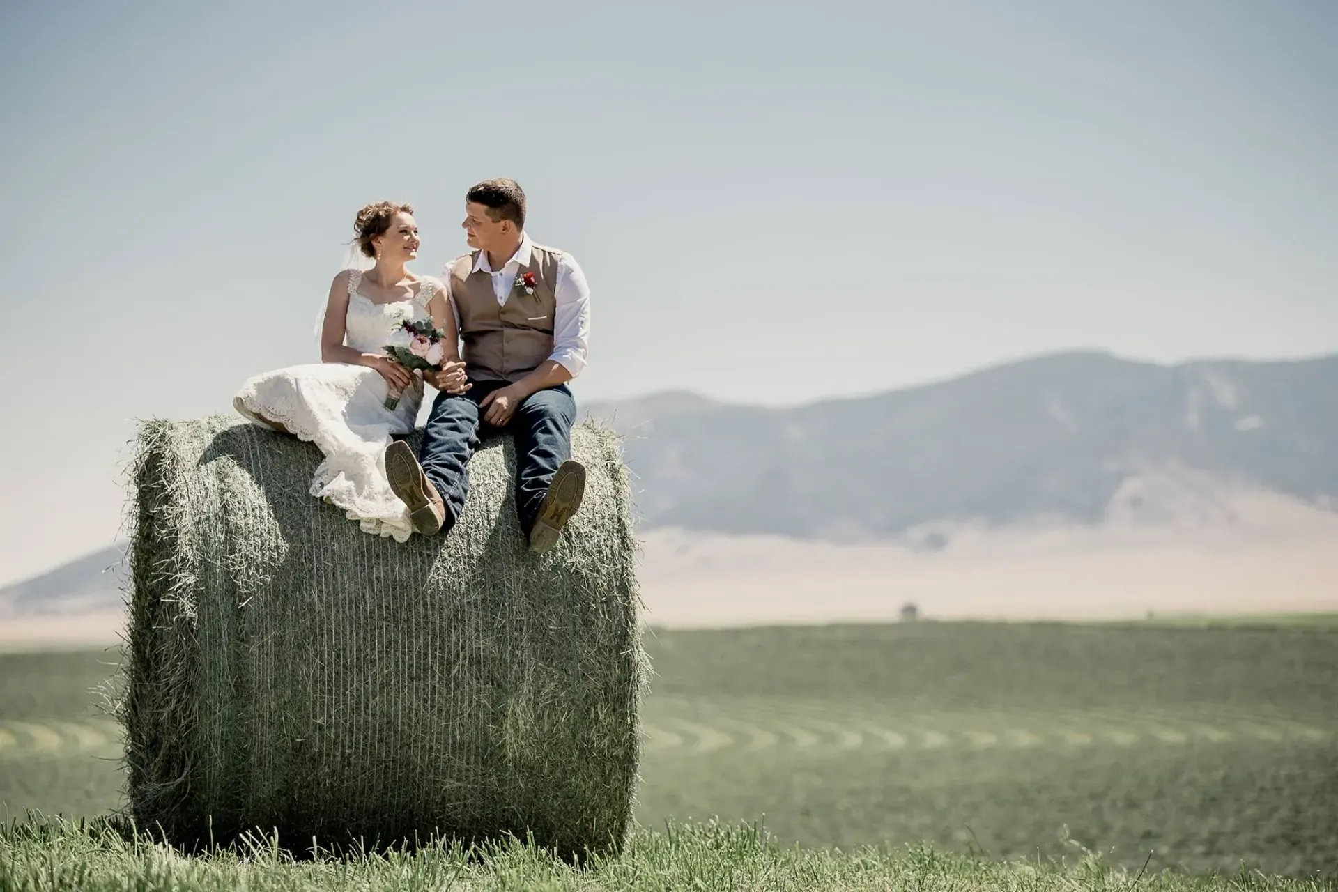 Two people sitting on a hay bale in a sunny field with mountains in the background
