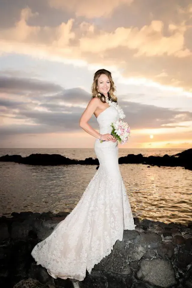 Bride in a white lace gown holding a bouquet on a rocky shore at sunset