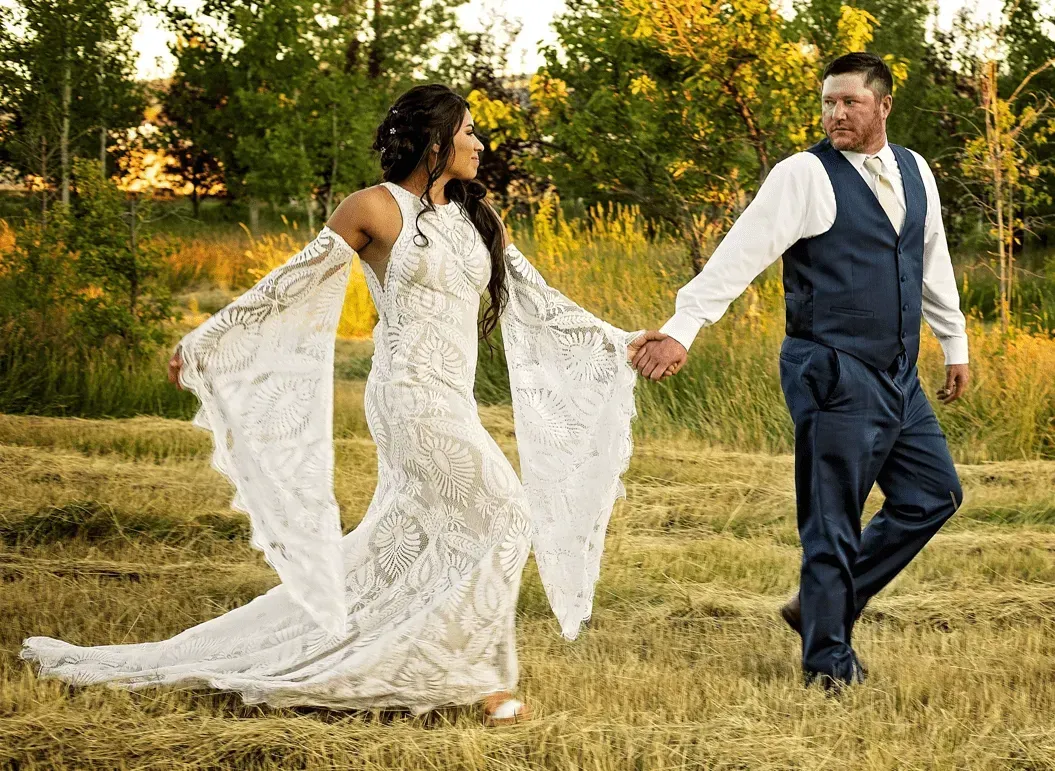 Couple in wedding attire holding hands and walking through a grassy field at sunset