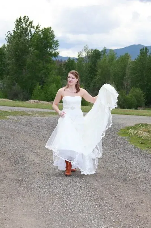 Person in a white dress holding the hem on a gravel path with trees and mountains in the background
