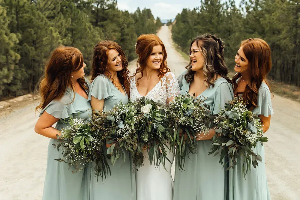 Five women in sage dresses holding greenery bouquets on a road surrounded by trees.