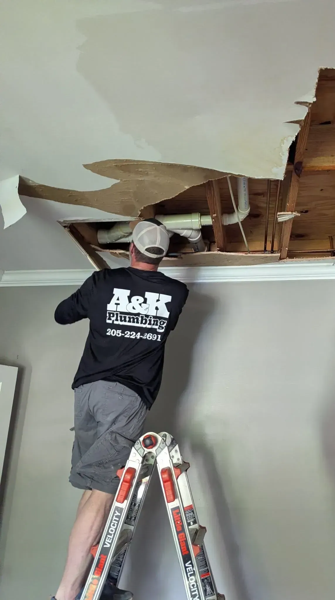 Worker on a ladder repairing a damaged ceiling with exposed wood and drywall.