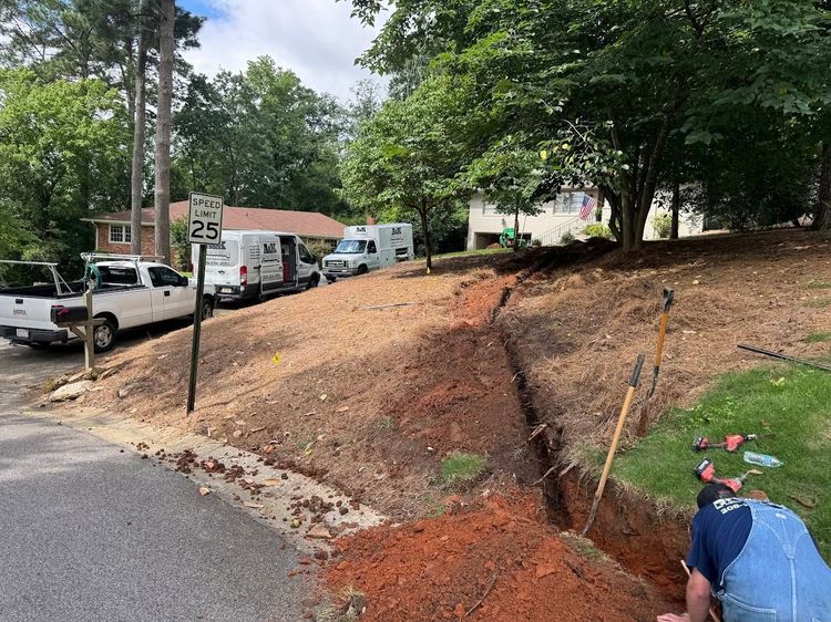 Construction worker inspecting a dug-out dirt slope beside a road and parked trucks in a wooded area