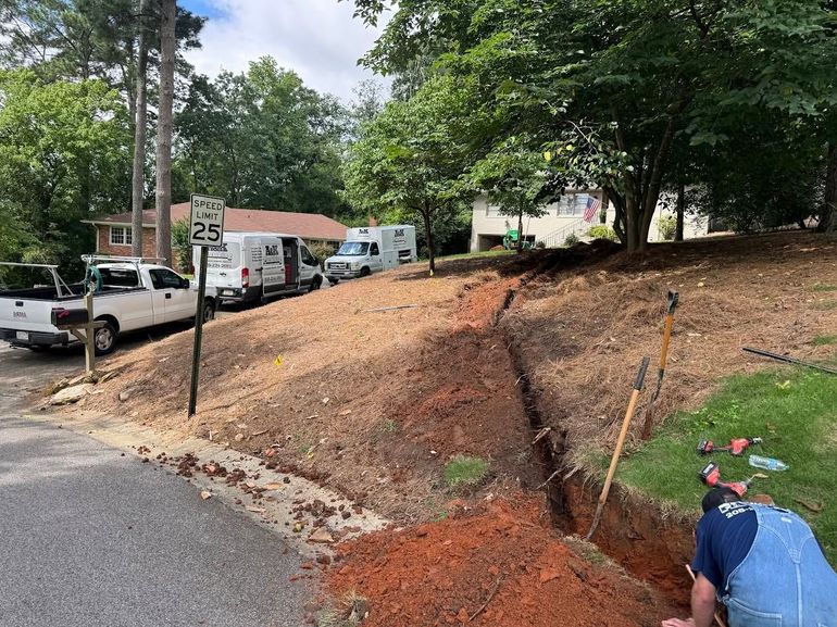 Construction worker inspecting a dug-out dirt slope beside a road and parked trucks in a wooded area